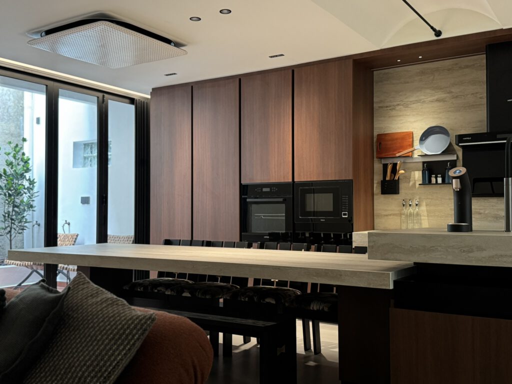 Modern open-plan kitchen with a long stone-topped island and bar seating, tall wood cabinetry with built-in oven and microwave, and a sink area with open shelves beside full-height glass doors.