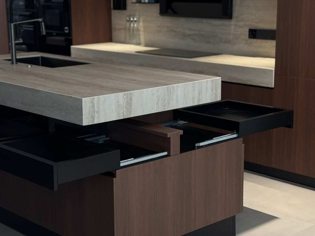 Close-up of a kitchen island with a thick sintered stone countertop and warm wood cabinetry, showing deep soft-close drawers pulled open with black interiors and smooth sliding mechanisms for cookware storage.