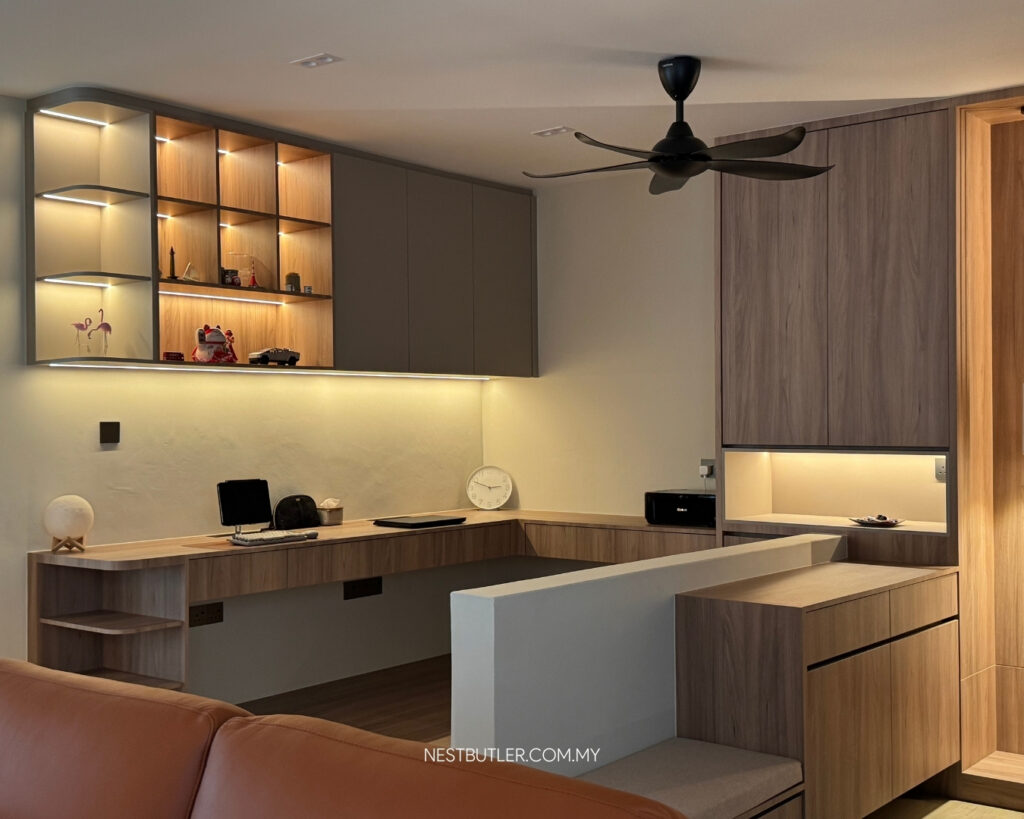 Modern built-in home office with warm wood cabinetry, LED-lit display shelves, an L-shaped desk, and a ceiling fan in a Malaysian living space.
